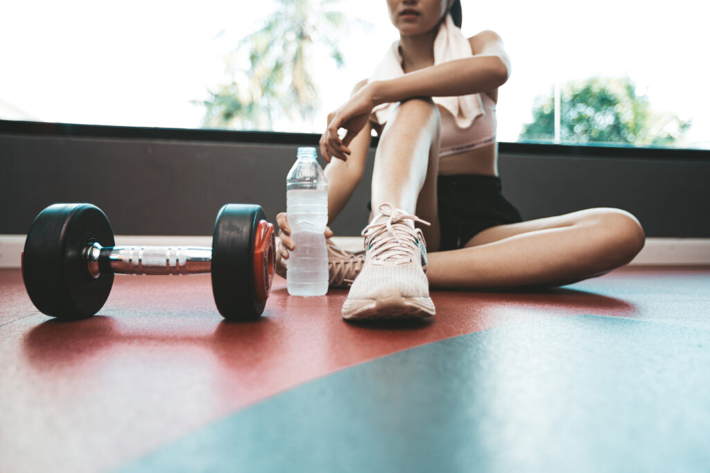 Mulher sentada e relaxada após o exercício. Há uma garrafa de água e halteres