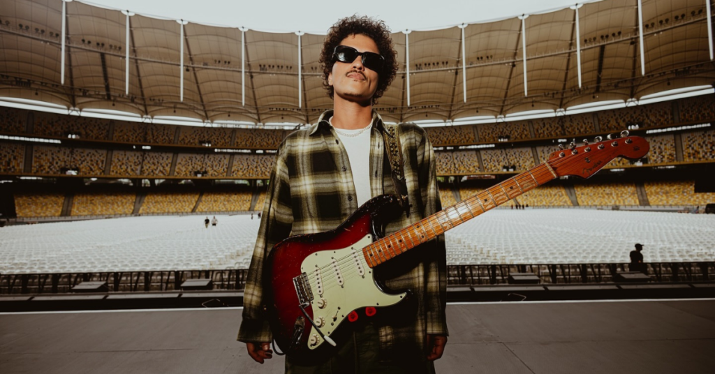 Bruno Mars com uma guitarra no palco de um estádio.