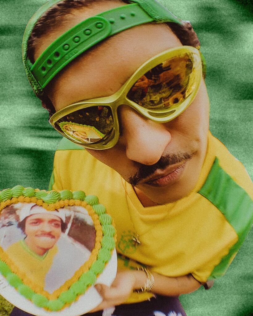 Bruno Mars vestido com uma camiseta verde e amarela, segurando um bolo de aniversário  em formato de coração com as cores do Brasil